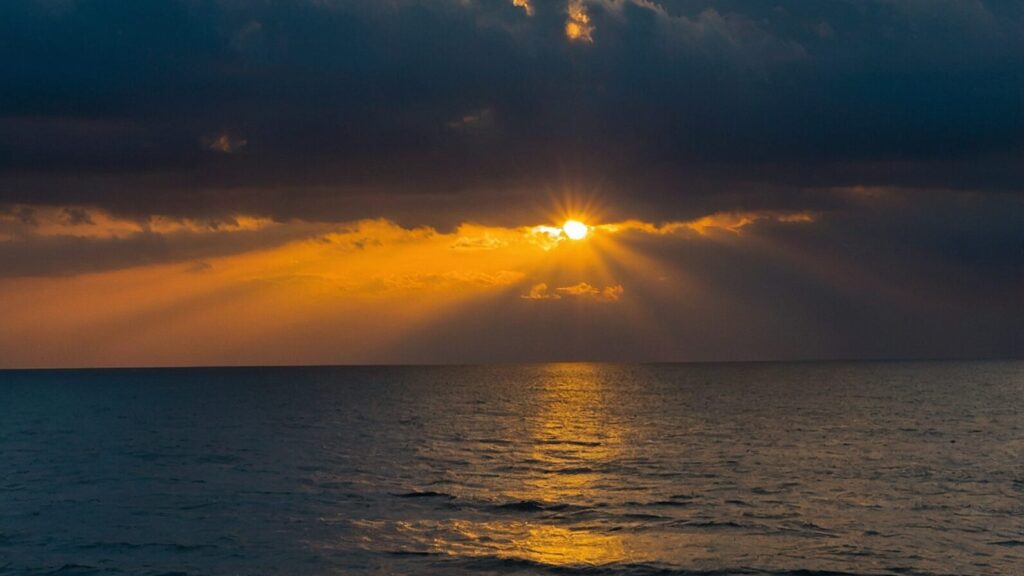 A scenic view of the ocean at sunset, with rays of light breaking through dark clouds.